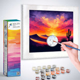Paint by numbers kit showing a hand painting a vibrant desert sky with a sunset, cacti, and rolling hills, labeled 