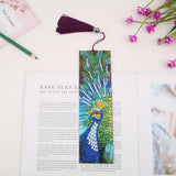 Colorful peacock design bookmark with a purple tassel placed on an open book with pink flowers and a green pencil nearby.