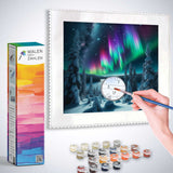 Paint by Numbers kit for Northern Lights in Secret Forest, showing a hand painting an outlined canvas with colorful paints and brush
