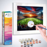 Malen nach Zahlen - Abendglühen im Stadion - Paint by numbers kit with a sunset stadium scene, a hand painting, and painting supplies.