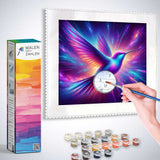 Guided diamond painting kit featuring a vibrant hummingbird in colorful purples and blues with a hand holding a brush ready to paint.